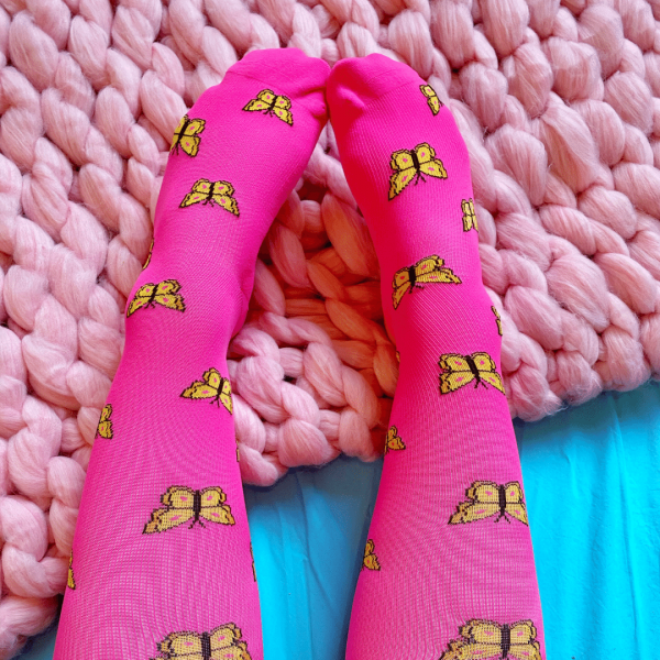 Bright magenta compression socks with yellow butterlies. The background is a pink knitted blanket with a blue sheet.