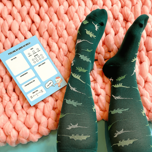 Dinosaur compression socks on a pink knitted blanket. A notebook is also lying on the blanket